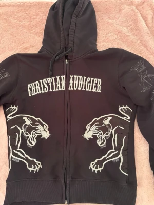 Christian Audigier svart hoodie - Svart hoodie med dragkedja från Christian Audigier. Stora vita tryck av pantrar på framsidan och broderad logga på ärmen. Huva med dragsko och detaljerade broderier ger en unik streetstyle-look.
