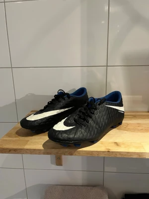 Nike Hypervenom svart/vit fotbollssko - Snygga Nike Hypervenom fotbollsskor i svart med vita detaljer och blå insida. Skorna har en texturerad ovandel för bättre bollkontroll och klassisk snörning. Perfekta för dig som vill sticka ut på planen och få grym känsla med bollen.