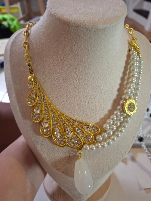 Guldigt halsband med pärlor och kristaller - Unikt halsband med en sida i guldfärgad metall dekorerad med gnistrande kristaller och en stor vit droppe. Andra sidan har dubbla rader vita pärlor och ett guldfärgat myntliknande hänge. Perfekt statement-piece för dig som vill sticka ut.