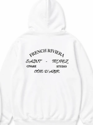 Vit hoodie från Civare Studio - Snygg vit hoodie från Civare Studio med tryck på ryggen där det står 'French Riviera', 'Saint-Tropez' och 'Côte d'Azur'. Framsidan har en liten svart logga och en klassisk känguruficka. Hoodien har dragsko i huvan och är gjord i mjukt material.