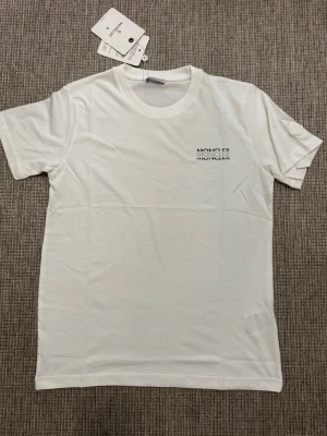 Vit t-shirt från Moncler - Vit t-shirt från Moncler med diskret logotyp på bröstet och ett emblem på ärmen. Klassisk rund halsringning och korta ärmar. Materialet är mjuk bomull som känns skönt mot huden. Perfekt för dig som gillar stilrena plagg med exklusiv känsla.