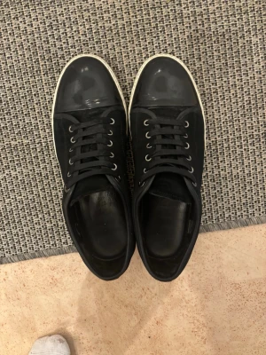 Svarta Lanvin sneakers i läder och textil - Snygga svarta sneakers från Lanvin med ovandel i både läder och textil. Skorna har rund tå, svarta skosnören och en vit sula med Lanvin-logga undertill. Tåpartiet är i blankt läder för en extra clean look. UK9