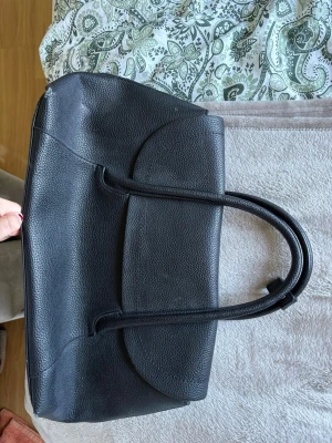 Svart klassisk handväska i skinn - Snygg svart handväska i slätt och lätt strukturerat skinn med två rejäla handtag. Väskan har en stilren och tidlös design med rundade kanter och rymligt huvudfack. Perfekt för dig som gillar minimalistisk och elegant stil. Slitage på väskan 