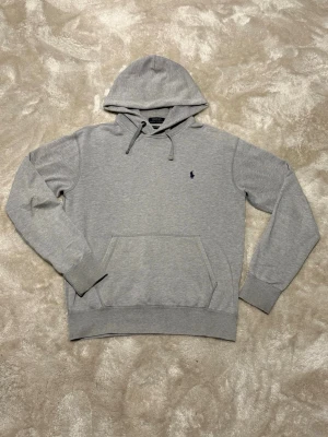 Polo Ralph Lauren hoodie - Säljer en snygg grå hoodie från Polo Ralph Lauren. Klassisk modell med liten marinblå logga framtill och mjuk insida. Hoodien är riktigt skön och passar bra. Den är i storlek S och är i väldigt bra skick. Den har endast en liten slitning vid ena armen men det syns knappt och är väldigt litet (sista bilden). Äkta Mått: Längd 63,5 cm Bröst 51 cm Armlängd 60 cm Axel 49 cm  Skickar snabbt!!📦🚚
