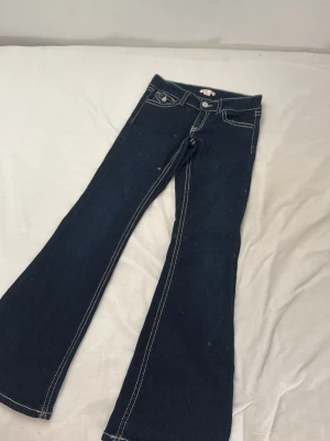 Mörkblå bootcut jeans från ginatricot - Mörkblå bootcut jeans från ginatricot med kontrasterande vita sömmar och klassisk femficksdesign. Jeansen har utsvängda ben och silverfärgade knappar. Perfekta för en trendig och avslappnad look.