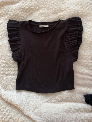 Svart topp med volangärmar från Zara - Snygg svart topp från Zara med ribbad struktur och volangdetaljer på axlarna. Modellen är ärmlös men har breda volangärmar som ger en cool och trendig look. Perfekt att styla med jeans eller kjol.