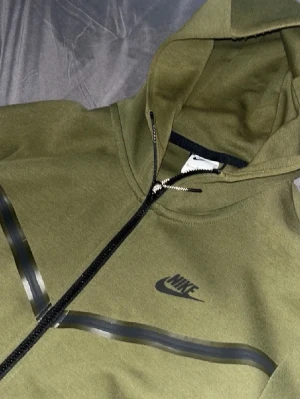 Olivgrön Nike hoodie med dragkedja och Byxor - Snygg olivgrön hoodie från Nike med svart logga på bröstet, dragkedja framtill och svarta snören i huvan. Hoodien har detaljer i form av svarta ränder och är tillverkad i mjukt bomullsmaterial. Perfekt för en avslappnad streetstyle-look.