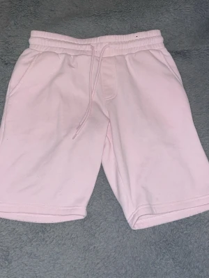 Ljusrosa t-shirt med vit logga - Säljer en ljusrosa t-shirt med en stor vit grafisk logga på bröstet. T-shirten har rund halsringning och korta ärmar. Materialet känns mjukt och bekvämt, perfekt för en avslappnad stil.