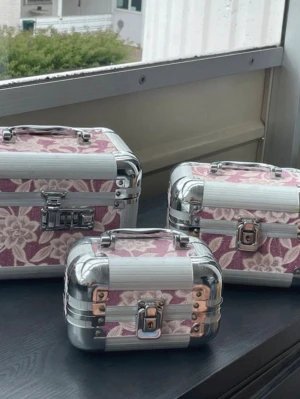 Blommiga smyckesboxar i metall - Tre smyckesboxar i olika storlekar med blommigt mönster i rosa och vitt. Boxarna har silverfärgade metallhörn, handtag och lås. Perfekta för att förvara smycken eller småprylar. Snygg design med stabil konstruktion och praktiska handtag.