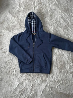 Marinblå burberry hoodie - Snygg marinblå hoodie med dragkedja och huva. Insidan av huvan och öppningen har ett rutigt foder i blått, vitt och rött. Hoodien har två fickor framtill och justerbara snören i huvan. Perfekt för en avslappnad och stilren look.