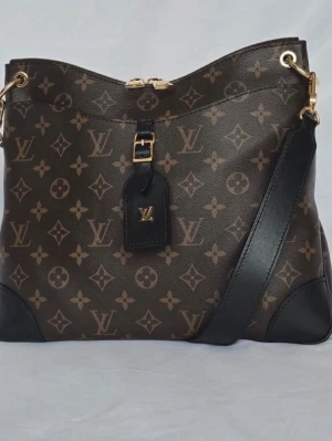 Louis Vuitton Monogram axelväska - Snygg axelväska från Louis Vuitton i klassiskt brunt monogrammönster med svarta läderdetaljer och guldfärgade metalldelar. Väskan har justerbar axelrem och ikonisk LV-tag framtill. Perfekt för dig som vill ha en lyxig och trendig accessoar. Dustbag kartong och kvitto medföljer 