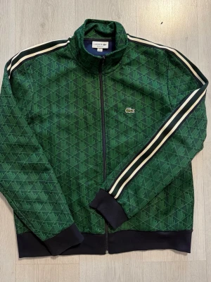 Grön ziptröja från Lacoste - Snygg grön ziptröja från Lacoste med diskret geometriskt mönster och vita ränder längs ärmarna. Tröjan har hög krage, muddar i svart och dragkedja framtill. Klassisk krokodil-logga på bröstet. Perfekt för en sportig och trendig look.