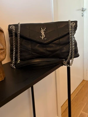 Svart YSL handväska i läder - Snygg svart handväska SL med quiltat mönster, silverfärgad Y S L -logga och kedjeremmar i metall. Väskan är tillverkad i mjukt äkta läder och har en klassisk kuvertformad design med lock. Aa***