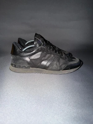 Svarta Valentino sneakers med nitar - Snygga svarta sneakers från Valentino med coola nitar på hälen och kamouflagemönster i mocka och läder. Skorna har lågt skaft, svarta skosnören och en chunky sula. Perfekta för dig som gillar streetstyle och exklusiva detaljer.