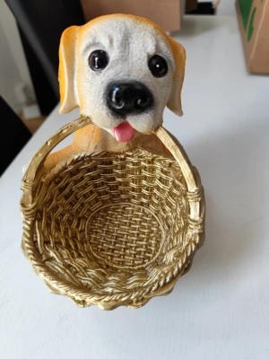Dekorativ hund med korg - Söt dekoration i form av en hund med en flätad guldfärgad korg. Hunden är ljusbrun och vit med stora svarta ögon och rosa tunga. Perfekt som prydnad eller förvaring av småsaker. Tillverkad i plast eller resin.