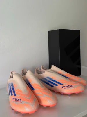 Adidas F50 Sparkfusion - Snygga Adidas F50 fotbollsskor i en fräsch kombination av ljusorange och beige med blå detaljer. Skorna har en strömlinjeformad design utan snörning och är tillverkade i syntetmaterial för bästa prestanda på planen. Perfekta för dig som vill sticka ut på matchen.