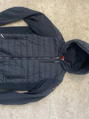 Svart vindjacka med huva - Snygg svart vindjacka från Parajumpers med quiltad kropp och släta ärmar. Jackan har huva, två dragkedjeförsedda bröstfickor och ribbade muddar. Märkespatch på ärmen och dragkedja framtill. Perfekt för blåsiga dagar.