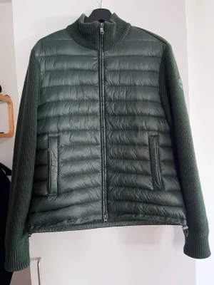 Grön hybrid jacka från Colmar - Stilren grön pufferjacka från Colmar med stickade ärmar och krage. Jackan har quiltad front och rygg, två dragkedjeförsedda fickor och en hög krage. Perfekt för kalla dagar och snygg detalj med Colmar-logga på ärmen.