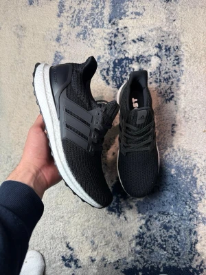 Adidas Ultraboost svarta sneakers - Svarta Adidas Ultraboost sneakers med stickad ovandel och klassiska tre ränder i plast på sidorna. Skorna har en vit Boost-mellansula och svart yttersula. Snygg och sportig design med snörning och låg profil. Perfekta för dig som gillar streetstyle och komfort. OBS! Ingen box ingår 