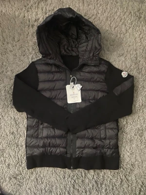 Svart pufferjacka med huva från Moncler - Snygg svart pufferjacka från Moncler med stickade ärmar och huva. Jackan har dragkedja framtill, ribbade muddar och Moncler-logga på ärmen. Perfekt för kalla dagar och riktigt skön att bära. Materialmix av dun och stickat ger en modern look.nfc funkar 