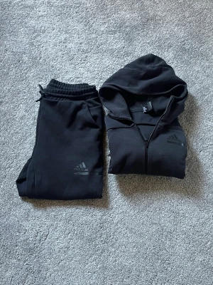 Adidas Mjukisset - Säljer ett svart mjukisset ifrån adidas. Hoodien har en klassisk huva och Adidas-logga på bröstet.  Mjukissbyxorna har snören och en logga på höger lår. Tveka inte på att skriva vid frågor, har svarar snabbt!