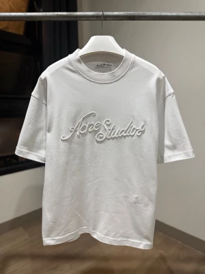 Vit Acne Studios t-shirt med brodyr - Vit t-shirt från Acne Studios med broderad logga framtill i samma färg som tyget. Klassisk rund halsringning och korta ärmar. Tillverkad i mjuk bomull för en clean och stilren look.