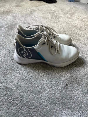 Vita golfskor från Footjoy  - Snygga vita sneakers från Footjoy med grå och blå detaljer på sidan samt logga. Skorna har lågt skaft, snörning och ovandel i mesh-liknande material för bra andningsförmåga. Perfekta för dig som gillar sportig och clean stil.