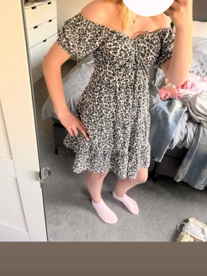 Blommig offshoulder klänning - Söt kort blommig klänning med offshoulder och volangkant. Klänningen har svartvitt blommönster, knyt framtill och är tillverkad i ett lätt bomullstyg. Perfekt för varma dagar och ger en romantisk känsla.