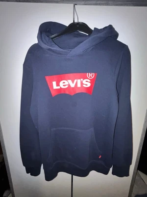 Mörkblå Levi's hoodie med logga - Snygg mörkblå hoodie från Levi's med stor röd och vit logga på bröstet. Klassisk känguruficka framtill och huva. Tillverkad i mjukt bomullsmaterial som är skönt att ha på sig. Perfekt för en avslappnad och trendig stil. Fett fläck på framsidan