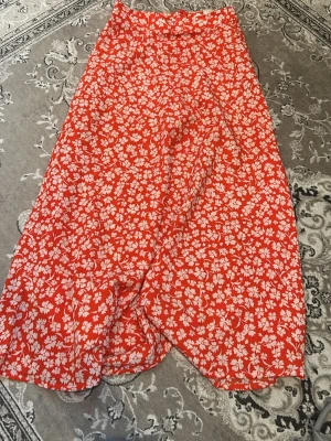 Orange blommig långkjol - Orange  långkjol med vitt blommönster. Kjolen har en lätt utsvängd form och är perfekt för dig som vill ha en färgstark och trendig look. Passar till både sneakers och sandaler. Materialet känns lätt och luftigt.