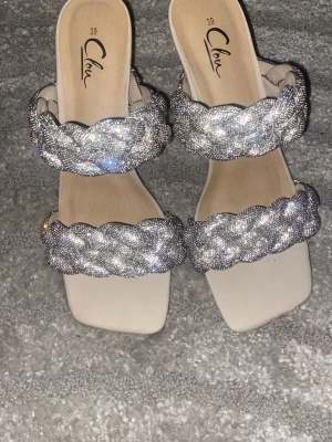 Glittriga sandaler med flätade band - Säljer ett par sandaler från Clou med två breda, flätade band täckta av silvriga diamanter . Sandalerna har fyrkantig tå och en ljusbeige sula. Perfekta för dig som vill sticka ut med extra bling och trendig design.