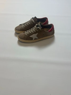 Beige Golden Goose Superstar sneakers storlek 41 - Snygga beige Golden Goose Superstar sneakers med vit stjärna på sidan och röd detalj vid hälen. Skorna har beige snörning, svart/vit GGDB-tagg på plösen. Skorna är i storlek 41