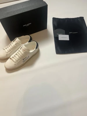 YSL vita sneakers storlek 41 - Snygga vita sneakers från YSL med svart detalj på hälen och handskriven logga på sidan. Skorna är låga och har klassisk snörning samt gummisula med fiskbensmönster. Tillverkade i läder för en clean och exklusiv look. Skorna har storlek 41 och är helt nya och aldrig burna