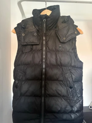 Svart dunväst från G-Star RAW - Svart dunväst från G-Star RAW med hög krage, avtagbar huva och flera fickor med knappstängning. Västen har en quiltad design och dragkedja framtill. Perfekt för lager-på-lager och streetwear-stil. Nypris ca 2000kr