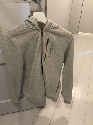 Ljusgrå hoodie från Sail Racing - Ljusgrå hoodie från Sail Racing med en olje fläck som tyvärr inte går bort och sönder i slutet vid handen som går att lagga. Och därmed är priset så lågt. Tillverkad i mjukt bomullsmaterial och har ribbade muddar vid ärmslut och nederkant. Klassisk huva och två sidofickor för extra funktion.