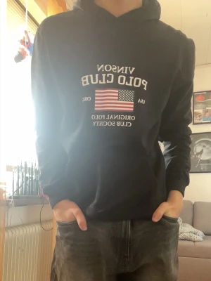 Svart hoodie från Vinson Polo Club - Svart hoodie från Vinson Polo Club med tryck av amerikansk flagga och vit text på bröstet. Klassisk känguruficka framtill och huva. Perfekt för en avslappnad och sportig stil.
