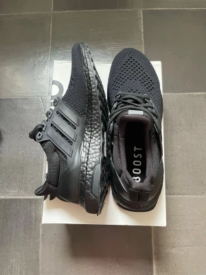 Adidas Ultra Boost sneakers - Snygga helsvarta Adidas Ultra Boost sneakers med stickad ovandel i mesh och klassiska tre ränder på sidan. Skorna har en responsiv Boost-sula och svart hälkappa för extra stöd. Perfekta för dig som gillar stilrena och bekväma sneakers.