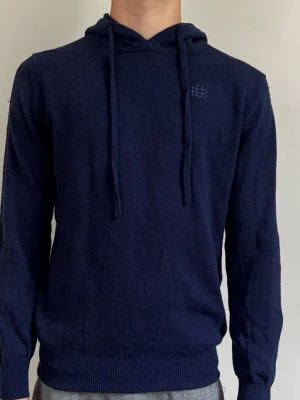 Mörkblå hoodie från Bvaldi - Mörkblå hoodie från Bvaldi med dragsko i huvan och diskret broderad logga på bröstet. Tillverkad i mjukt stickat material som känns skönt mot huden. Perfekt för dig som gillar en enkel och stilren look.