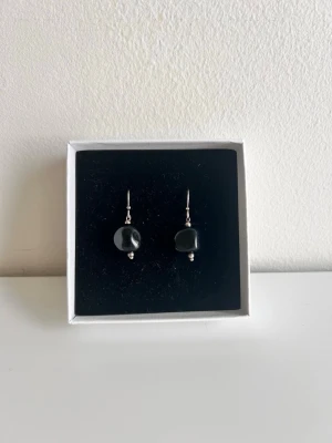 Svarta örhängen med stenpärlor - Snygga sterling silver örhängen med svarta, oregelbundna stenpärlor och silverfärgade detaljer. Hängande modell med enkel krok, perfekta för att ge en stilren touch till din look. Kommer i en elegant ask.