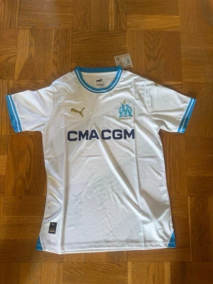 Vit Marseille matchtröja Puma - Snygg vit matchtröja från Olympique de Marseille med blå detaljer och klubbmärke på bröstet. Puma-logga i guld och sponsortryck 'CMA CGM' framtill. Tillverkad i lätt och ventilerande material, perfekt för fotboll eller träning.
