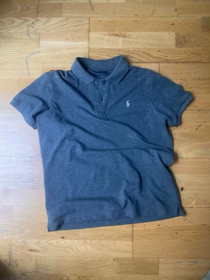 Mörkgrå pikétröja från Ralph Lauren - Klassisk mörkgrå pikétröja från Ralph Lauren med korta ärmar och vit broderad logga på bröstet. Tröjan har krage och knappar framtill, perfekt för en clean och stilren look.