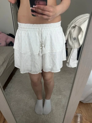 Vit volangkjol med broderidetaljer - Söt vit kjol med volanger och små broderade detaljer i färg. Kjolen har resår i midjan och dekorativa tofsar framtill. Lätt och luftig modell som passar perfekt till sommaren.