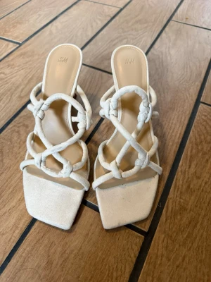 Beige klackade sandaler från H&M - Snygga beige sandaler med klack från H&M. De har trendiga knutna remmar i tyg och fyrkantig tå. Perfekta för att lyfta din outfit med en modern touch. Klacken är medelhög och ger både stil och comfort. Helt nya och oanvända i storlek 40 men passar en 39 då de är små i storlek. Sulan nästan 27 cm och klacken 5 cm.