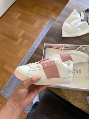 Valentino Garavani vita sneakers - Snygga vita sneakers från Valentino Garavani med en bred rosa rem över sidan och klassiska snörningar. Skorna har en chunky sula och är tillverkade i läder för en lyxig känsla. Perfekta för dig som vill sticka ut med stilrena detaljer.