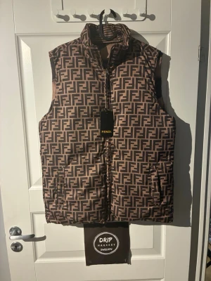 Fendi väst brun  - Snygg brun dunväst i storlek large från Fendi med ikoniskt FF-mönster över hela västen. som man kan vända in och ut. Västen har hög krage, dragkedja framtill och två fickor på sidorna. Perfekt för dig som vill sticka ut med en lyxig och trendig look.