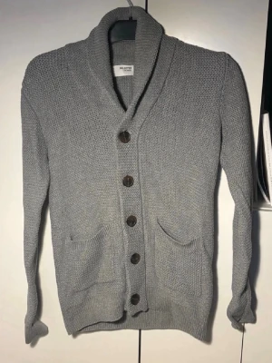 Selected homme cardigan  - Snygg grå stickad kofta från Selected med stora bruna knappar och två praktiska fickor framtill. Koftan har en bred krage och långärmad design, perfekt för lager-på-lager. Materialet är mjukt och strukturerat för en cool look.