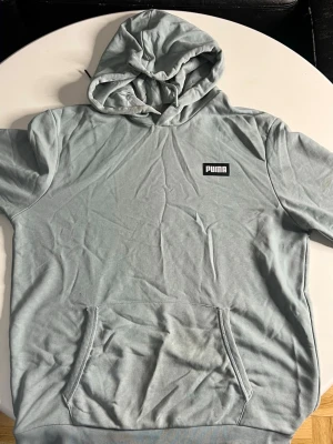 Ljusblå hoodie från Puma - Snygg ljusblå hoodie från Puma med klassisk huva och magficka. Tröjan har korta ärmar och Puma-logga på bröstet. Tillverkad i mjukt bomullsmaterial som känns skönt mot huden. Perfekt för en avslappnad och sportig stil.