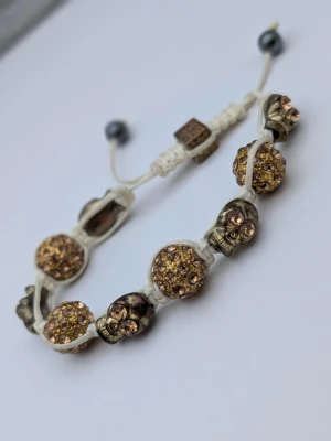 Armband med döskallar av mässing och glittriga pärlor - Coolt armband med vit justerbar snodd, dekorerat med guldfärgade glittriga pärlor och metallfärgade döskalleberlocker. Armbandet har en unik design med detaljerade döskallar och gnistrande pärlor som ger en edgy och trendig look.