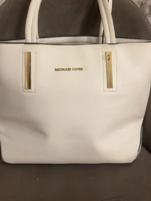 Vit handväska från Michael Kors - Stilren vit handväska från Michael Kors med guldfärgade detaljer och dubbla handtag. Väskan har en rak, fyrkantig form och är tillverkad i ett tåligt konstläder. Baksidan har en praktisk ficka med dragkedja. Perfekt för dig som gillar klassisk och elegant design.