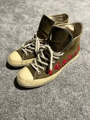 Converse x CDG Play olivgröna highs - Säljer ett par Converse x Comme des Garçons Play sneakers i olivgrönt canvasmaterial med vita sulor och snören. Sidan pryds av flera röda hjärtan med ögon, en ikonisk detalj från CDG Play. Klassisk high top-modell med gummitå och metallöljetter.